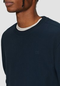 Navyblauwe geribbelde sweatshirt met een ronde hals en subtiele QS-logo op de borst. Gelaagd met een witte onderhemd en gemaakt van textuurstof.