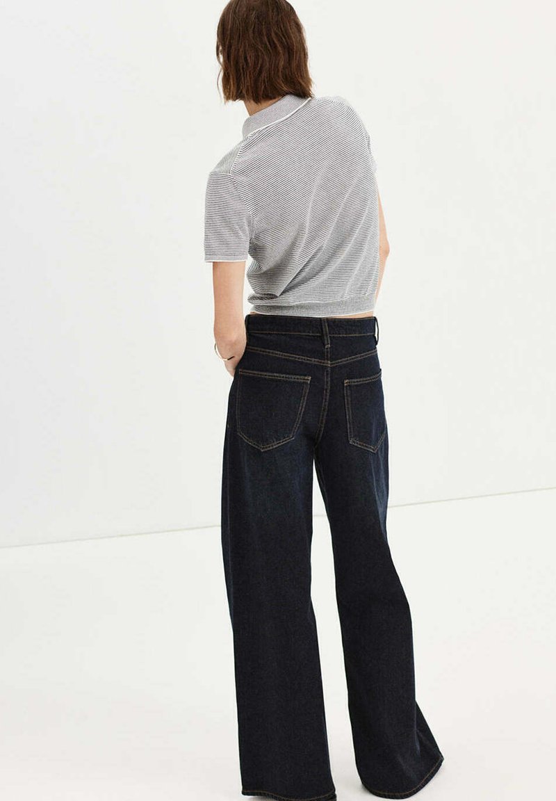 Personne aux cheveux courts bruns portant un haut gris rayé à manches courtes et un jean large foncé, debout les mains dans les poches, de dos.