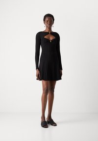 Vestido preto canelado com mangas longas, um decote recortado e um detalhe decorativo em dourado. Saia godê com pregas verticais, combinada com mules pretas.