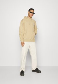 Sudadera beige con un bolsillo frontal y un sutil detalle de logo, combinada con pantalones de pierna ancha de color claro y zapatos negros. Los accesorios incluyen gafas de sol.