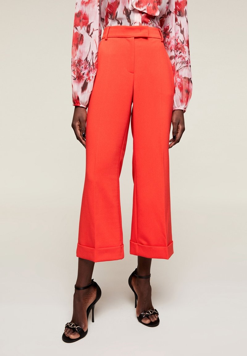 Kvinna som bär ljusröda vida cropped byxor, blommig långärmad blus och svarta högklackade sandaler med kedjedetaljer.