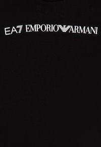 Črna bombažna majica s poudarjenim belim logotipom: "EA7 EMPORIO ARMANI," z linearni poudarki na črkah.