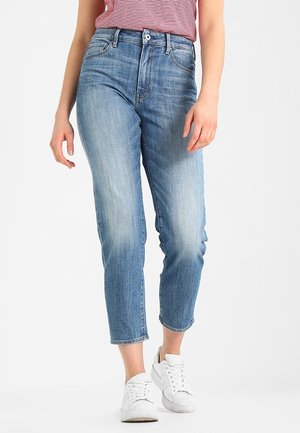 Kvinde iført lyseblå cropped jeans, hvide sneakers og en rød- og hvidstribet indstukket skjorte, stående mod en ensartet baggrund.