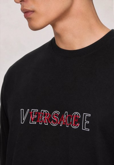 Млад човек, носещ черна тениска с наслагван червен и бял контур "VERSACE" текст през гърдите.