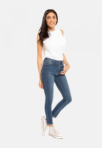Jean skinny bleu à taille haute, doté de passants pour ceinture et de poches avant, associé à un top blanc sans manches et des baskets blanches.