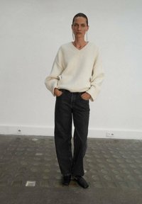 Suéter de punto color crema con escote en V, combinado con pantalones de mezclilla negros de pierna ancha de talle alto y zapatos negros tipo slip-on. Fondo escaso.