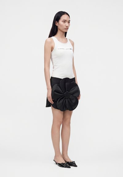 Femme portant un débardeur blanc avec un texte et une jupe noire avec un grand détail de fleur en tissu, assortie à des chaussures noires sans lacets.