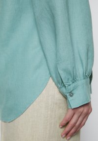 Una camicia a maniche lunghe di un azzurro chiaro con una consistenza morbida e polsini con bottoni, abbinata a un pantalone di lino di colore chiaro.