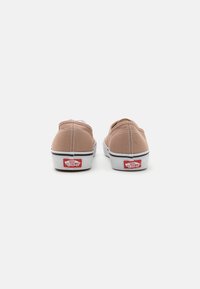 Paire de baskets slip-on Vans beige vues de dos avec des semelles en caoutchouc blanches et des logos rouges "Vans Off The Wall" sur un fond blanc uni.