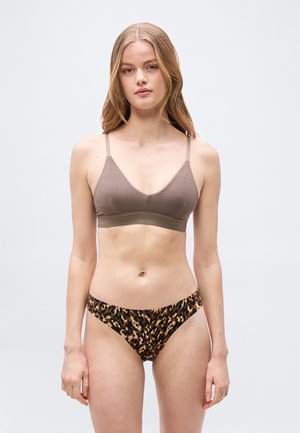 Νεαρή γυναίκα στέκεται με το πρόσωπο προς τα εμπρός, φορόντας ένα σουτιέν ταουπέ με λεπτές τιράντες και μαγιό σλιπ σε σχέδιο λεοπάρ, μπροστά σε ένα απλό ανοιχτόχρωμο φόντο.