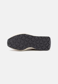 Semelle extérieure en caoutchouc noir avec une adhérence à motifs, présentant un intermédiaire beige pâle et un logo en relief sur le talon.