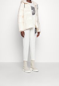 Puffig, ljusbeige jacka med huva och blixtlås fram. Kombinerad med vita byxor och beige vriststövlar, med en tryckt sweatshirt under.