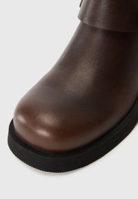 Stivale alla caviglia in pelle marrone con punta rotonda, superficie testurizzata e suola in gomma nera. Presenta una finitura liscia e un design semplice.