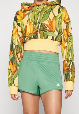 Person iført grønne Adidas sportsshorts og en gul cropped hættetrøje med stort orange og grønt tropisk blomsterprint.