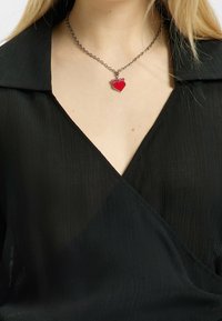 Collier en chaîne argentée avec un pendentif en forme de cœur rouge. Le pendentif a une finition brillante, posé sur un haut en tissu noir texturé.