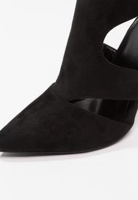 Chaussure à talon haut en daim noir pointue avec un design découpé près de la voûte plantaire et une semelle intérieure en cuir lisse.