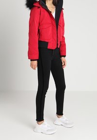 Červená zkrácená bunda s kožíškem na kapuci, dvěma předními kapsami, spárovaná s černými úzkými kalhotami s postranními pruhy a bílými teniskami.