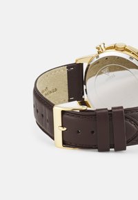 Cinturino in pelle marrone con fibbia e dettagli in tono oro. Cassa rotonda in acciaio inossidabile con finitura lucida, dotata di un fondello in vetro.