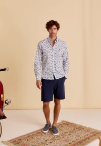 Knopenblouse met kleurrijk bloemenpatroon, lange mouwen en een witte basis. Gecombineerd met marineblauwe shorts en grijze schoenen op een geblokte vloerkleed.