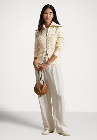 Kremasto pleten pulover z visokim ovratnikom, nošen čez lahkotno majico in široke bele hlače. V roki drži okroglo rjavo usnjeno torbico.