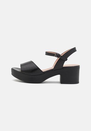 Sandale plateforme en cuir noir avec bride de cheville réglable et talon bloc, présentée sur un fond gris clair uni.
