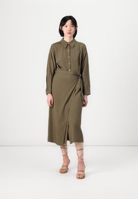 Olive zelené šaty košeľového strihu s golierom, dlhými rukávmi a prekrývajúcim dizajnom. Obsahuje gombíky a predný rozparok; skombinované s béžovými sandálmi na opätku.