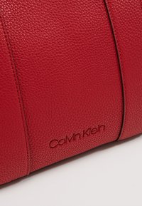 Gros plan sur un sac en cuir rouge texturé avec des coutures verticales et le logo Calvin Klein embossé près du centre en bas.