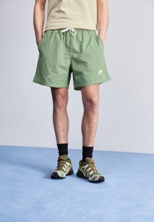 Zielone bawełniane szorty z białym sznurkiem i logo Nike. Połączone z zielonymi maskującymi trampkami i czarnymi skarpetkami za kostkę na jasnoniebieskiej podłodze.