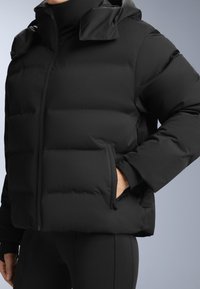 Crna puf jakna opuštenog kroja, s visokim ovratnikom, kvadratnim dijelovima i džepovima na zip. Glatka, mat tekstura.