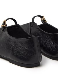 Zapatos negros de cuero texturizado con parte trasera baja y pequeña correa con hebilla. Presentan una suela plana y un forro interior suave.