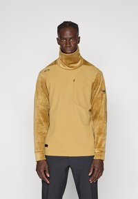 Sudadera de color mostaza con cuello alto, mangas de terciopelo texturizado, un bolsillo en el pecho y acentos de logo en el hombro.