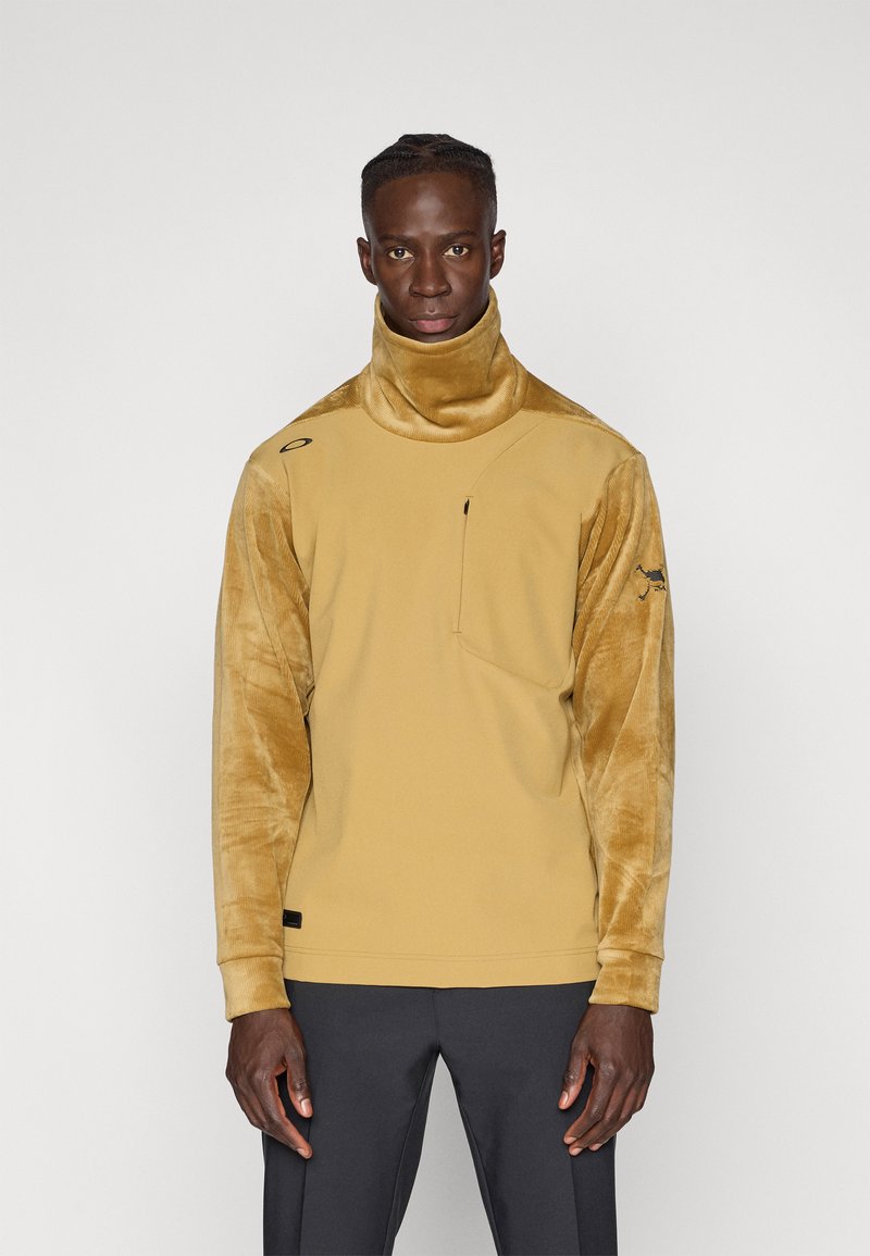 Sudadera de color mostaza con cuello alto, mangas de terciopelo texturizado, un bolsillo en el pecho y acentos de logo en el hombro.