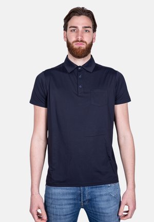 Uomo con capelli castani e barba, che indossa una polo blu scuro e jeans blu, in piedi davanti a uno sfondo chiaro e uniforme.