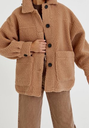 Person wearing a tan, textured teddy coat with black buttons and large front pockets, paired with matching brown pants.