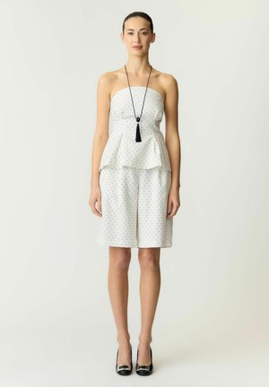 Vrouw gekleed in een strapless witte peplum-jurk met zwarte stippen, zwarte hakken en een lange zwarte kwastketting, die recht vooruit staat.