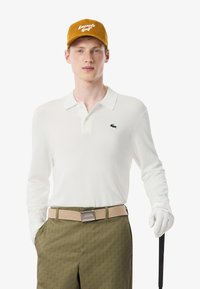 Polo bianco a maniche lunghe con logo verde, abbinato a pantaloni verdi a fantasia e una cintura beige. Un cappellino di velluto a coste giallo completa l'outfit.