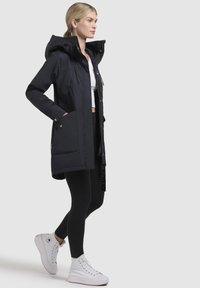 Black padded jacket with a detachable hood, front zip, and snap pockets, paired with black leggings and white high-top sneakers.