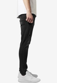 Černé džínsy s úzkým střihem a opranými detaily, stylizované s bílým tričkem a bílými teniskami, zobrazené z profilu.