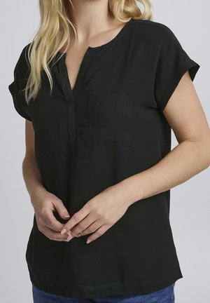 Woman with blonde hair wearing a short-sleeve black blouse with a front pocket and partial button placket, hands clasped at waist.