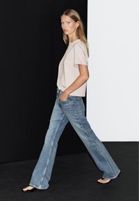 Beige T-Shirt mit kurzen Ärmeln, kombiniert mit hellblauen weit geschnittenen Jeans, die ein klassisches Fünf-Taschen-Design und einen faded Look aufweisen.