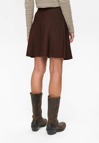 Femme portant une jupe marron midi, des bottes en cuir marron et une chemise à manches longues rayée, montrée de dos sur un fond blanc.