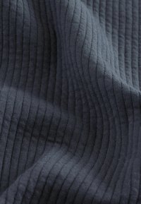 Dunkelgrauer Stoff mit einer strukturierten, gerippten Oberfläche. Das Muster zeigt abwechselnd erhabene und flache Linien, die Tiefe und Dimension erzeugen.