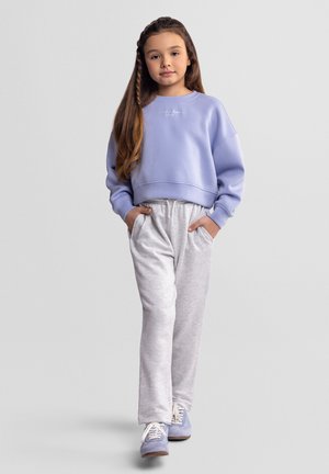 Noor tüdruk pikkade juustega, riietatud lilla dressipluusi, helehallide dressipükste ja lilla tossudega, seisab käed taskutes.