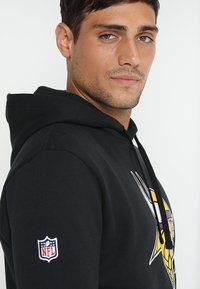 Sudadera negra con un gráfico en el frente que presenta a un vikingo. Incluye una capucha con cordones y un parche del logo de la NFL en la manga derecha. Textura suave.