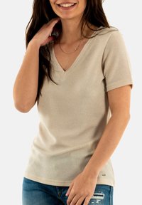 Top beige à col en V et manches courtes, fabriqué à partir d'un tissu tricoté texturé, avec une légère brillance. Le modèle l'associe à un jean en denim déchiré.