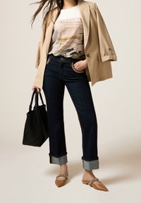 Woman wearing beige blazer over printed blouse, dark cuffed jeans with chain detail, tan pointed flats with embellishments, holding black woven bag.