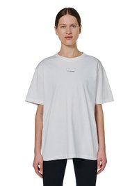 Frau mit braunen Haaren trägt ein lockeres weißes T-Shirt mit dem Schriftzug "HALO" und schwarze Hose, steht vor einem einfarbigen weißen Hintergrund.