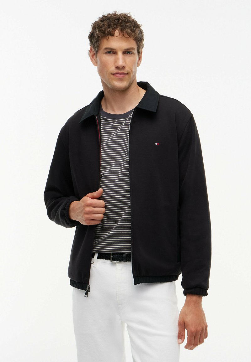 Young man wearing a black zip-up jacket over a black and white striped shirt with white pants standing against a plain white background.