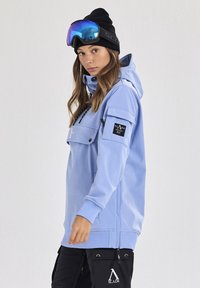 Helleblaue Skihand mit einer Fronttasche, verstellbarem Kapuze und schwarzen Akzenten. Getragen mit schwarzen Hosen, die Reißverschluss-Taschen haben.