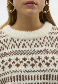 Strickpullover mit cremefarbener Basis und braunen geometrischen Mustern. Verfügt über einen gerippten Halsausschnitt und eine weiche Textur. Goldene Ohrringe mit Perlenakzent.